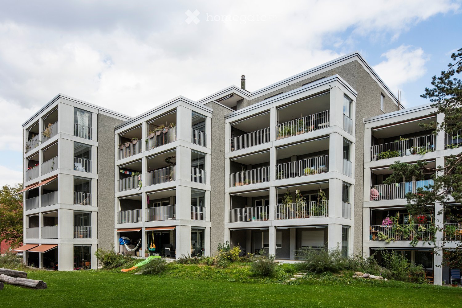 Modern multi-story apartment building with balconies, large windows, and greenery in the foreground.