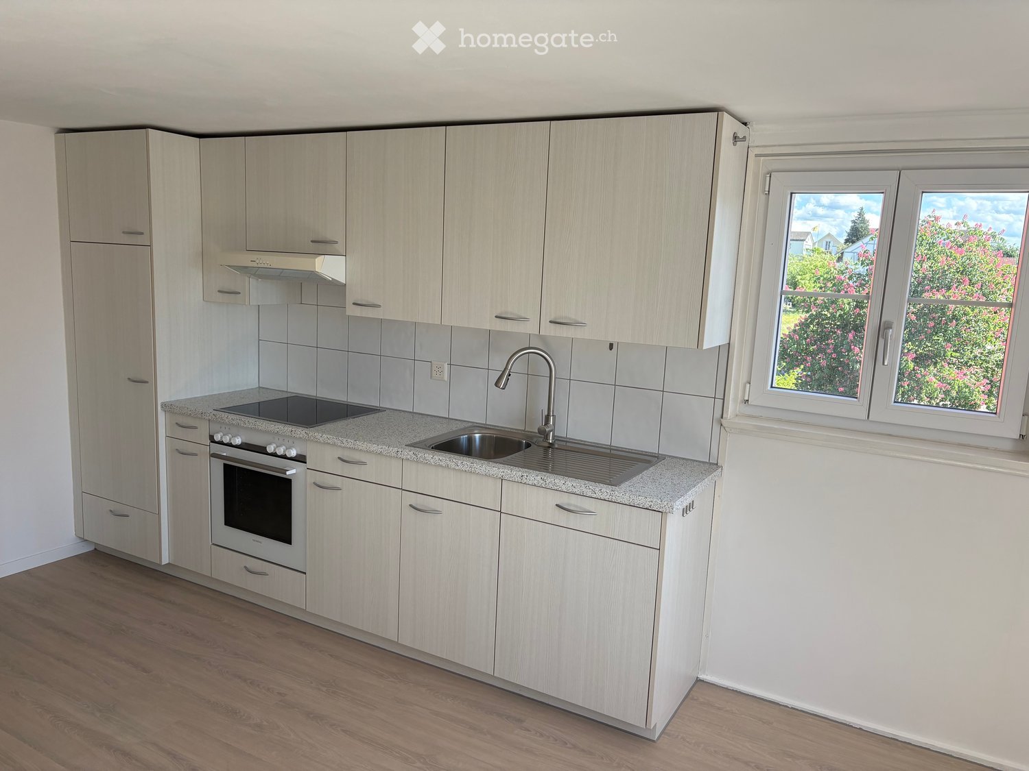 Modern kitchen with light wood cabinets, granite countertop, stainless steel sink and faucet, built-in oven, and range hood. Large windows offer a view of greenery outside.
