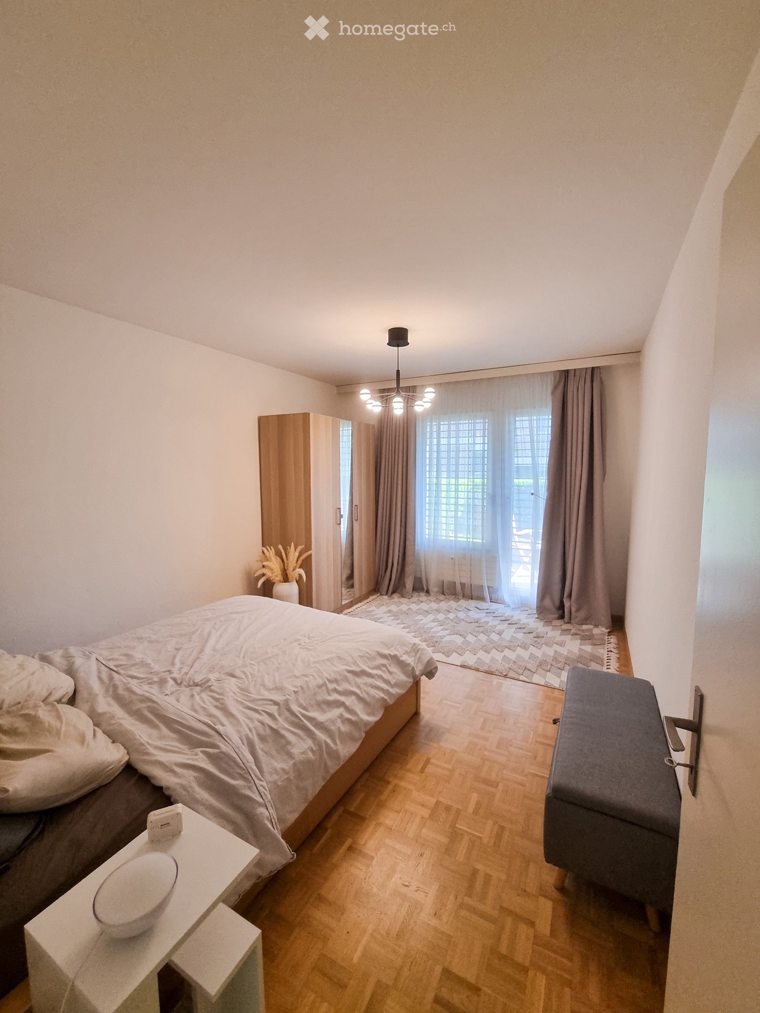 A modern bedroom with a large bed, nightstand, chair, wardrobe, and a view through a large window with curtains.