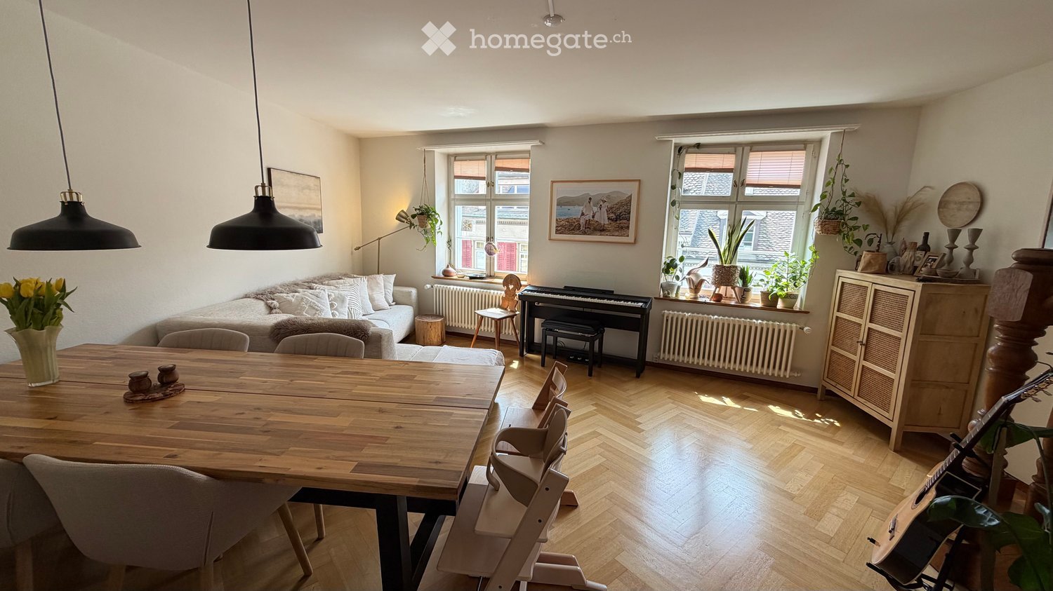 Bright living and dining area with a sofa, dining table, piano, and various decorative items including potted plants and artwork.