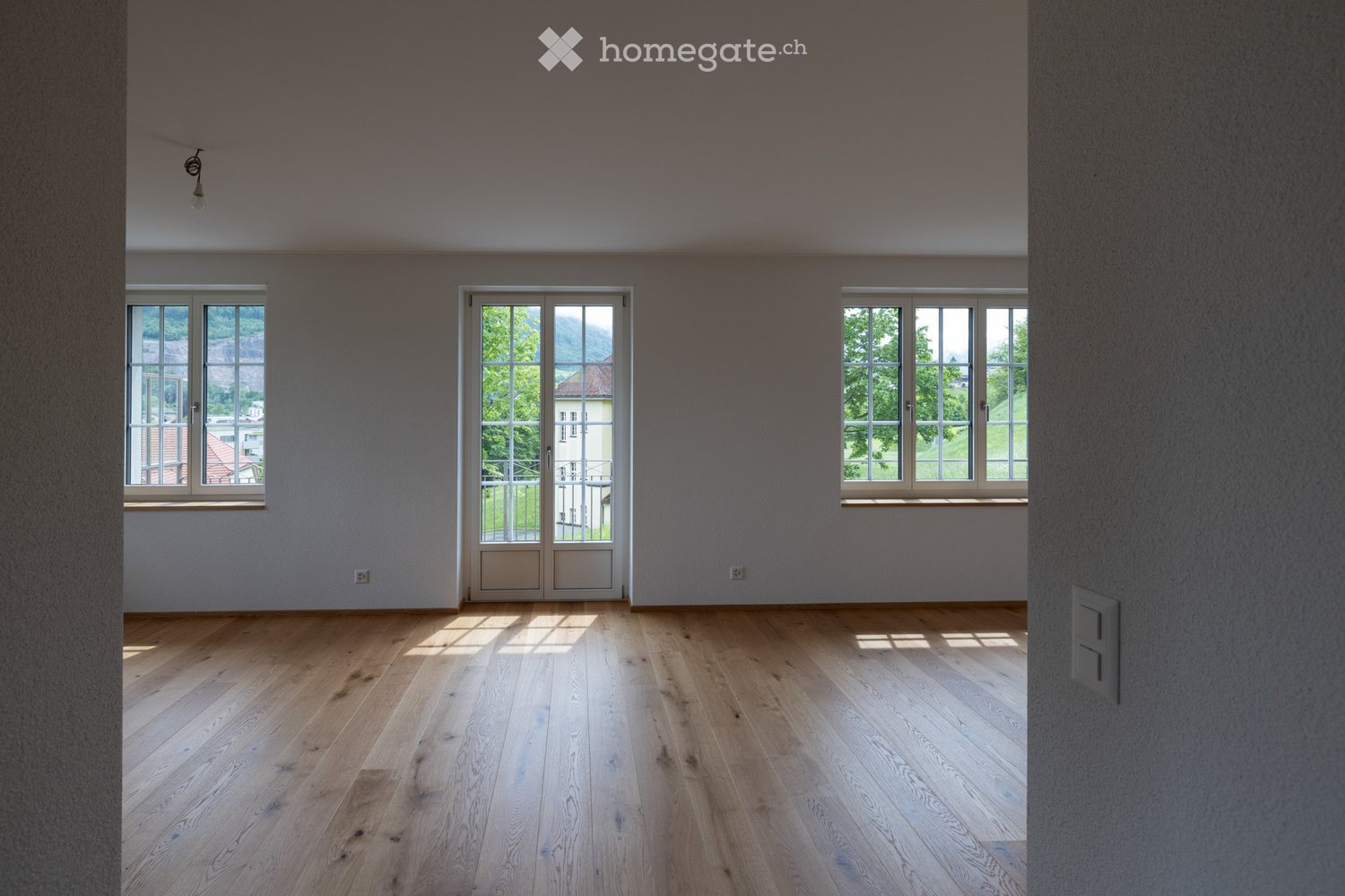 Empty room with light wooden flooring, large windows, and a door leading to a balcony.