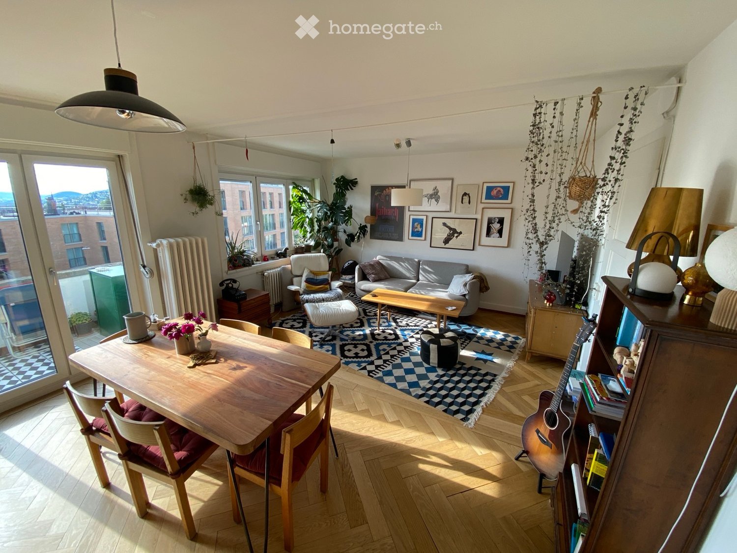 Bright living room with wooden flooring, large windows, a sofa, coffee table, dining table with chairs, guitar, bookshelf, and decorative items.
