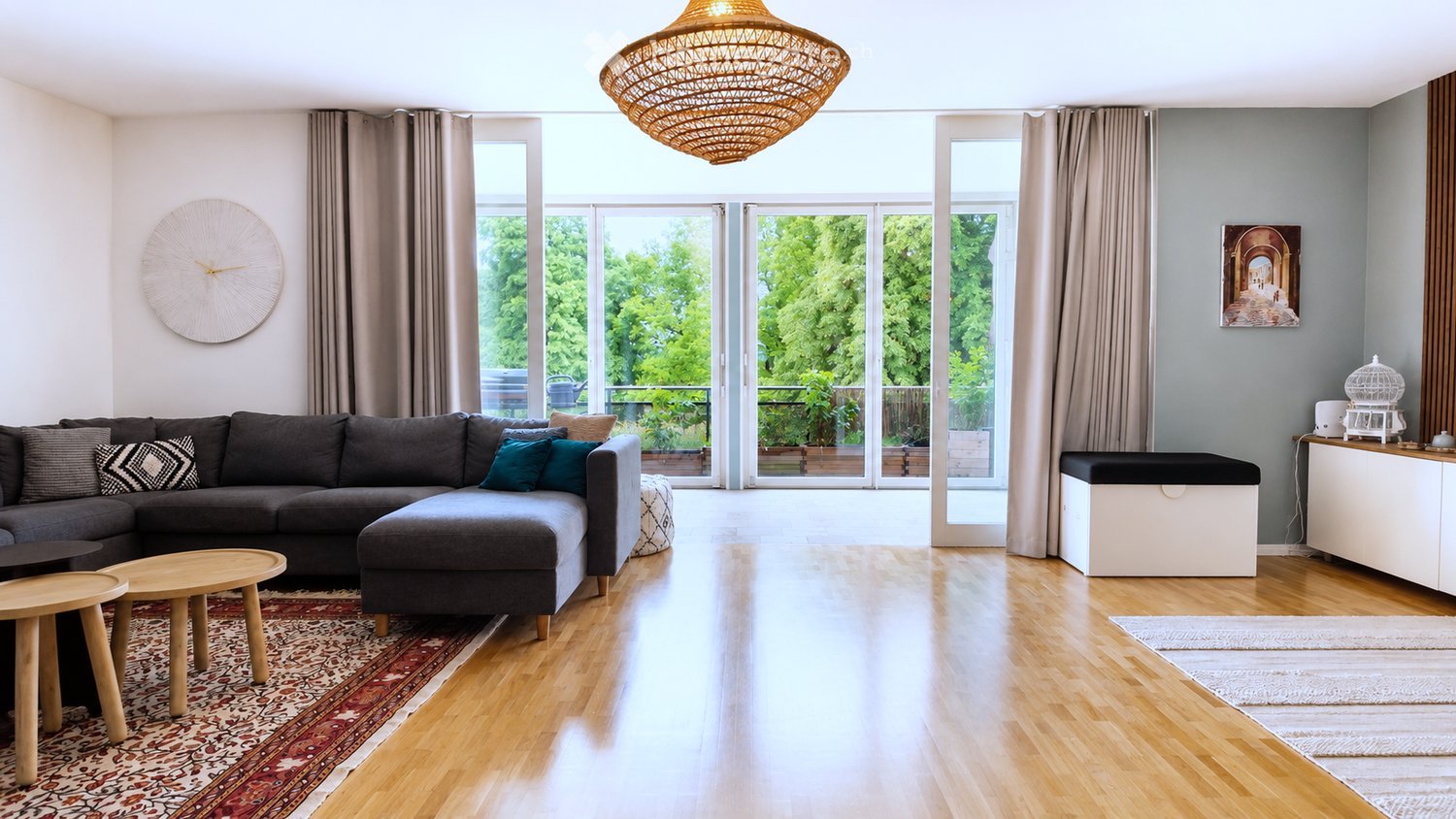 Modern living room with a grey sectional sofa, wooden coffee table, ottoman, and side tables. Features large windows with curtains, a woven hanging light fixture, and a view of greenery outside.