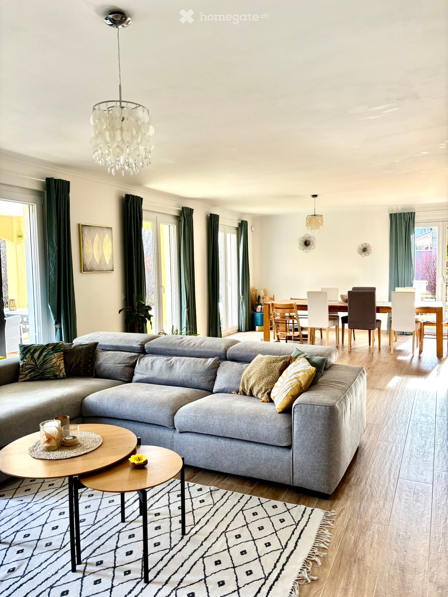 Modern living room with grey sectional sofa, wooden coffee tables, dining area with chairs and table, chandelier, and large windows with green curtains.