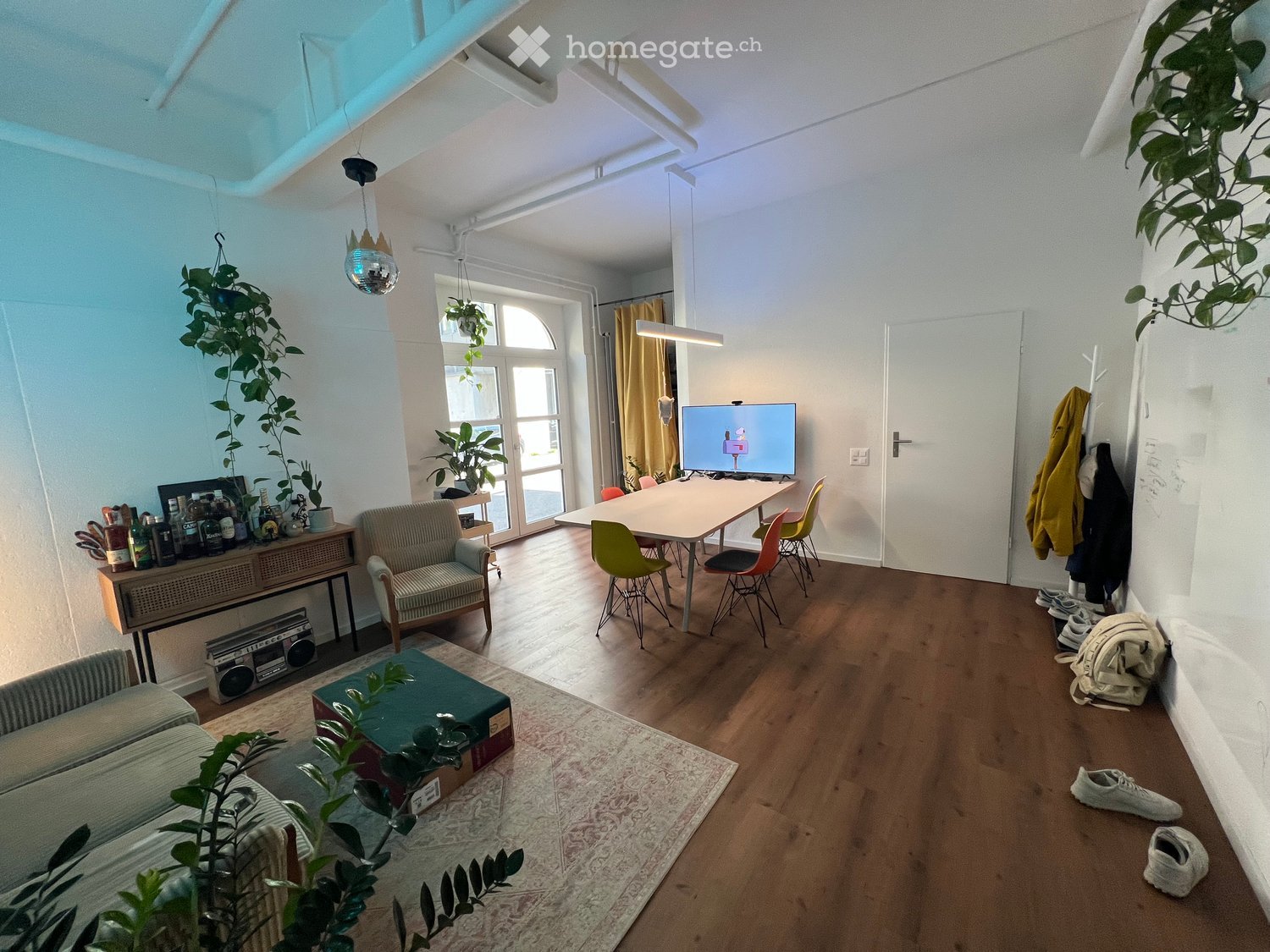 A modern, well-lit living room with wooden flooring, a large window, and a mix of seating and dining areas. Features include a sofa, armchair, coffee table, dining table with chairs, a television, and several plants.