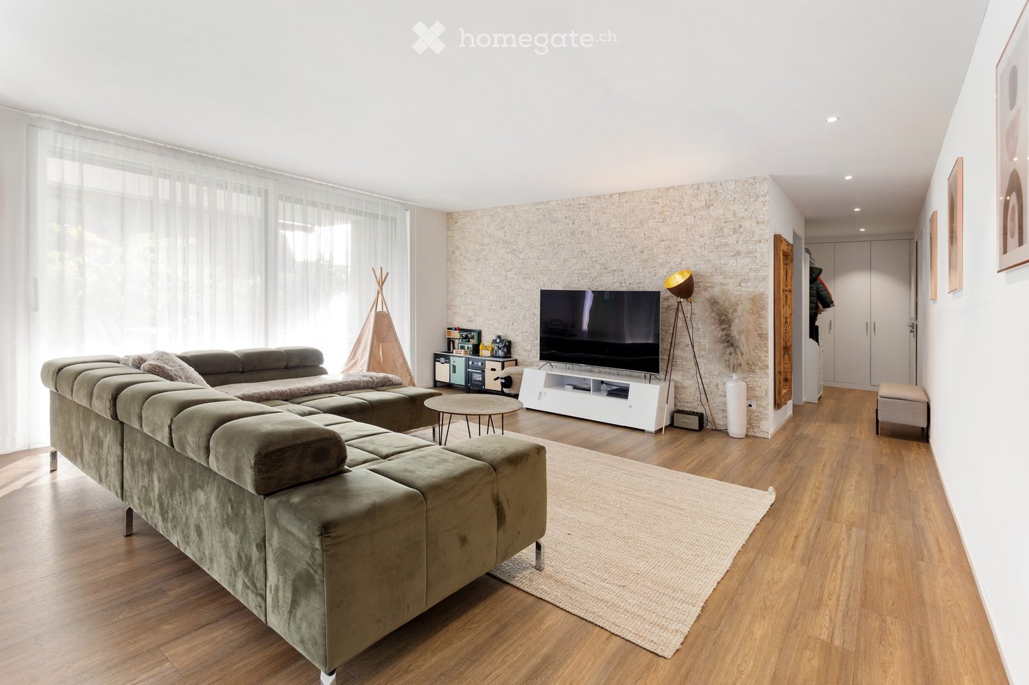 Modern living room with a sectional sofa, coffee table, television, floor lamp, side table, and decorative vase. The room features wooden flooring, large windows with sheer curtains, and a textured accent wall.