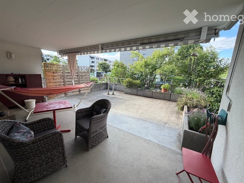 Balcony with outdoor furniture including wicker chairs, a red table, and a hammock, surrounded by greenery and a view of the city.