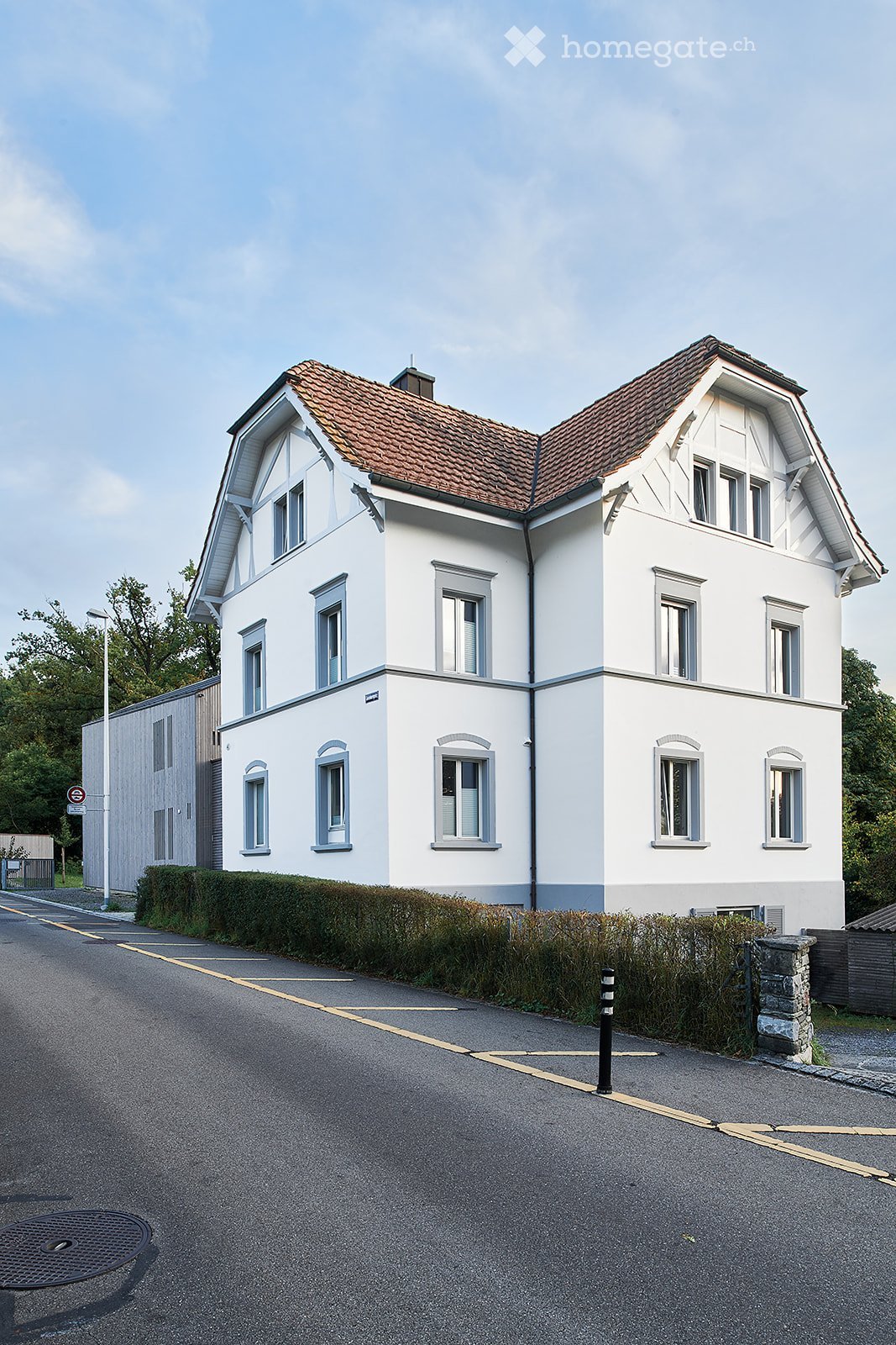 Two-story white house with a red-tiled roof, multiple windows, and a well-maintained hedge in front.