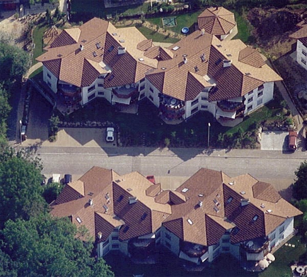Aerial view of a large residential complex with multiple buildings, brown tiled roofs, balconies, and surrounding greenery. A swimming pool and parking area are visible.