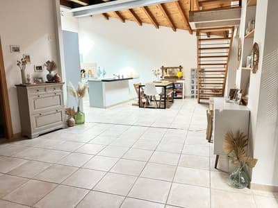 Bright, open-plan living and dining area with white tiled flooring, exposed wooden beams, and modern kitchen island. Features include a dresser, dining table with chairs, and decorative vases.
