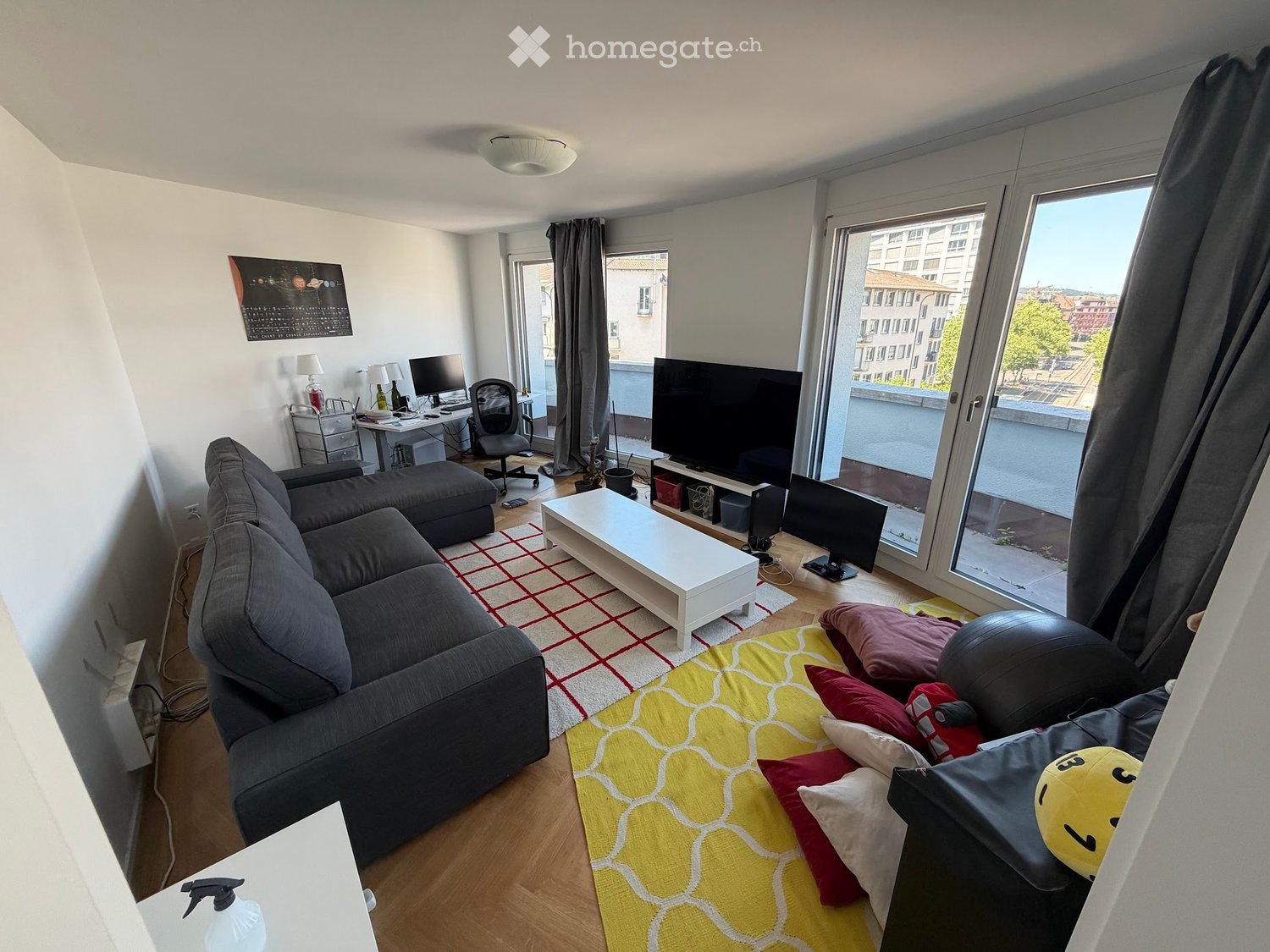 Modern living room with a grey sectional sofa, white coffee table, desk with computer, and large windows offering a city view.