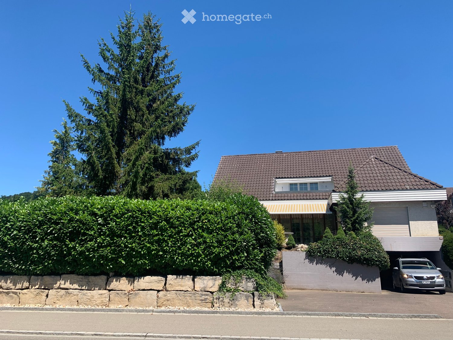 Two-story house with a tiled roof, garage, and surrounding greenery including a large tree and shrubs.
