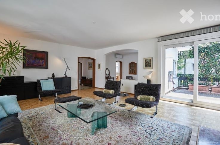 Modern living room with large windows and sliding glass doors leading to a balcony. Features a dark-colored sofa, glass coffee table, armchairs, and decorative items.