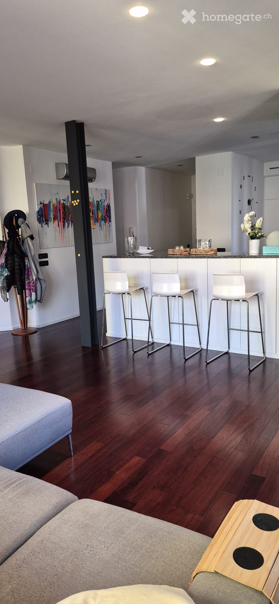Modern living room with dark wooden flooring, white bar area with stools, grey sofa, and decorative items including a vase and painting.