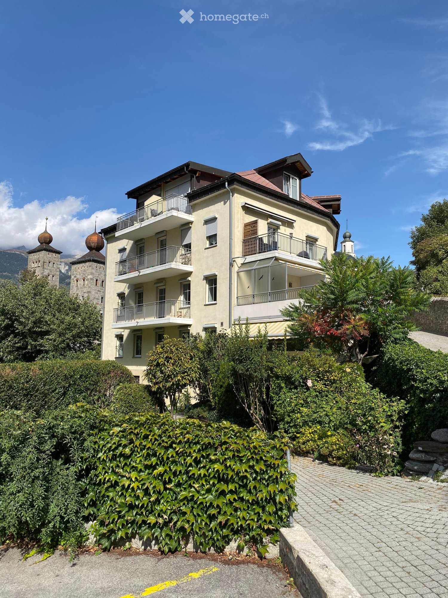 A multi-story residential building with balconies, surrounded by greenery and a stone pathway.