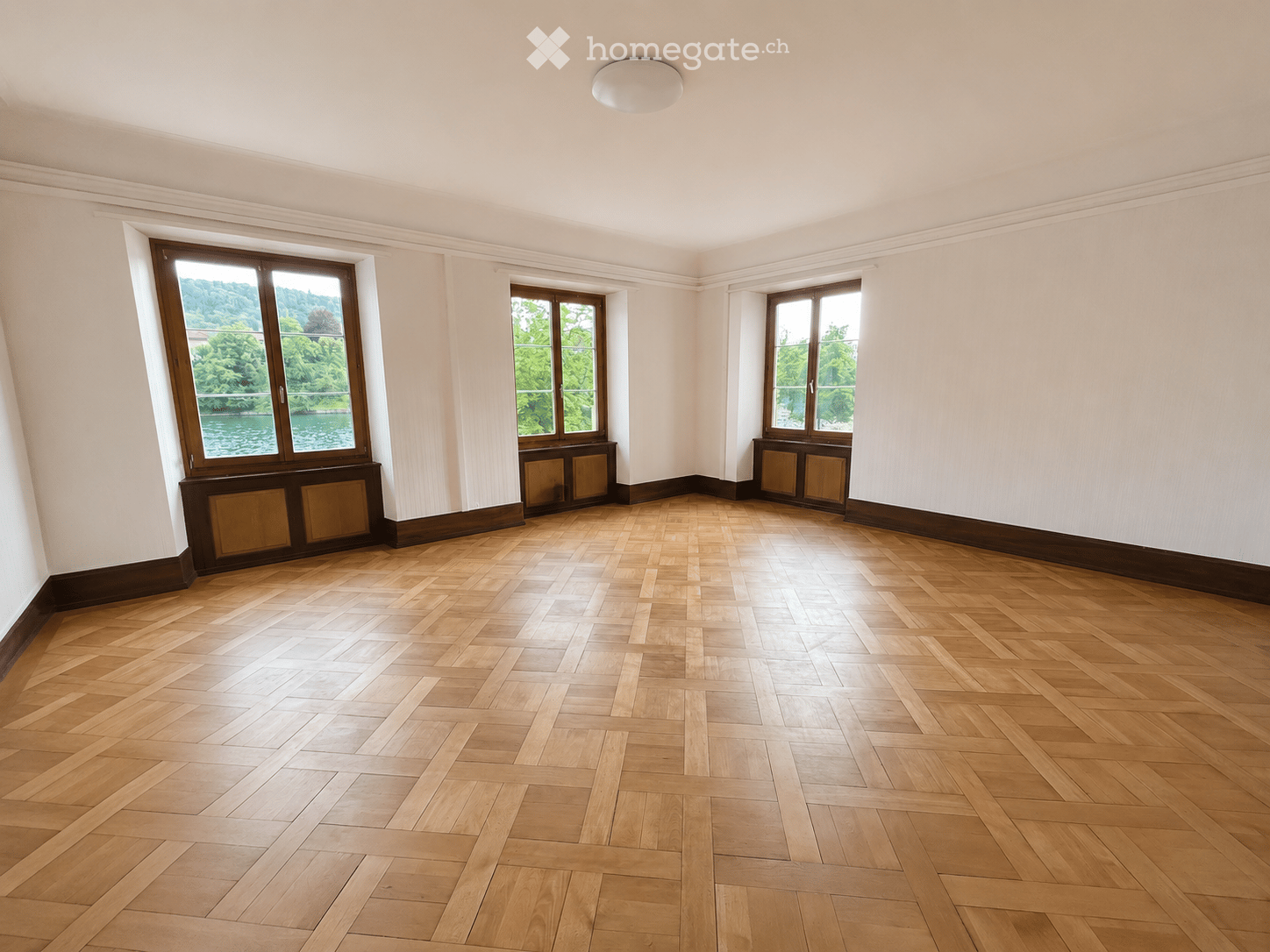 Empty room with wooden herringbone flooring, white walls, and large windows offering a scenic view of a river and greenery.