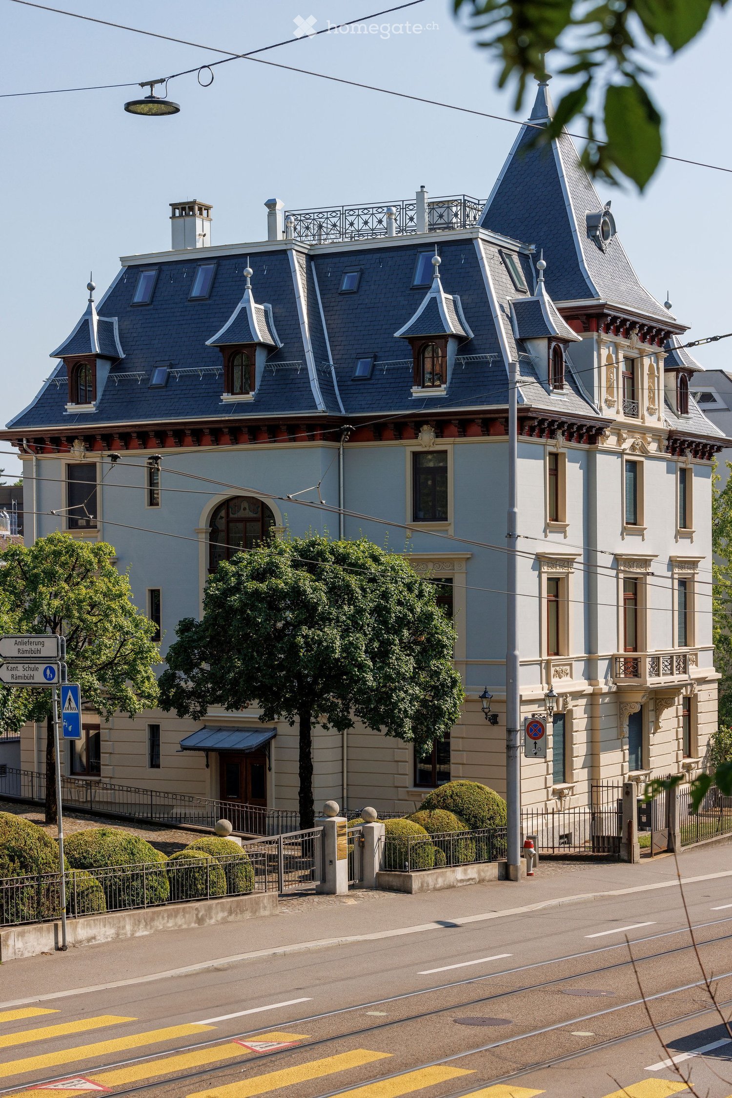 A large, ornate building with a steep, multi-pitched roof and decorative turrets. The facade is light blue with white trim and features arched windows and balconies. The building is surrounded by a low fence and well-maintained shrubs.