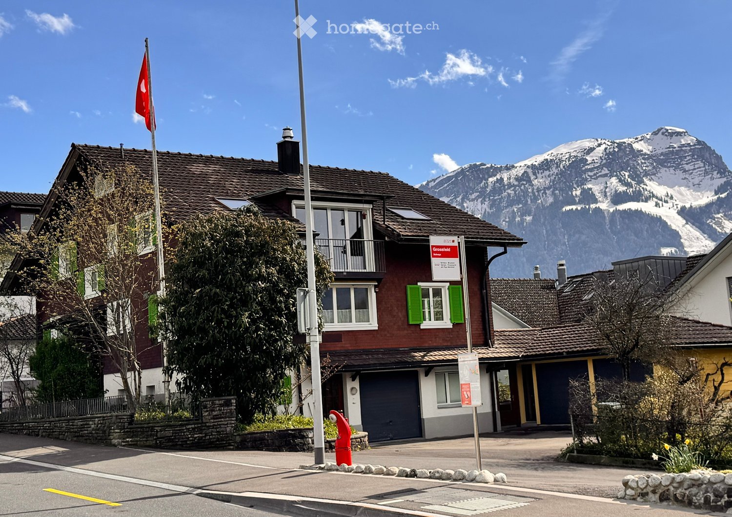 A two-story house with a red exterior, green shutters, and a balcony, situated in a picturesque village with snow-capped mountains in the background.