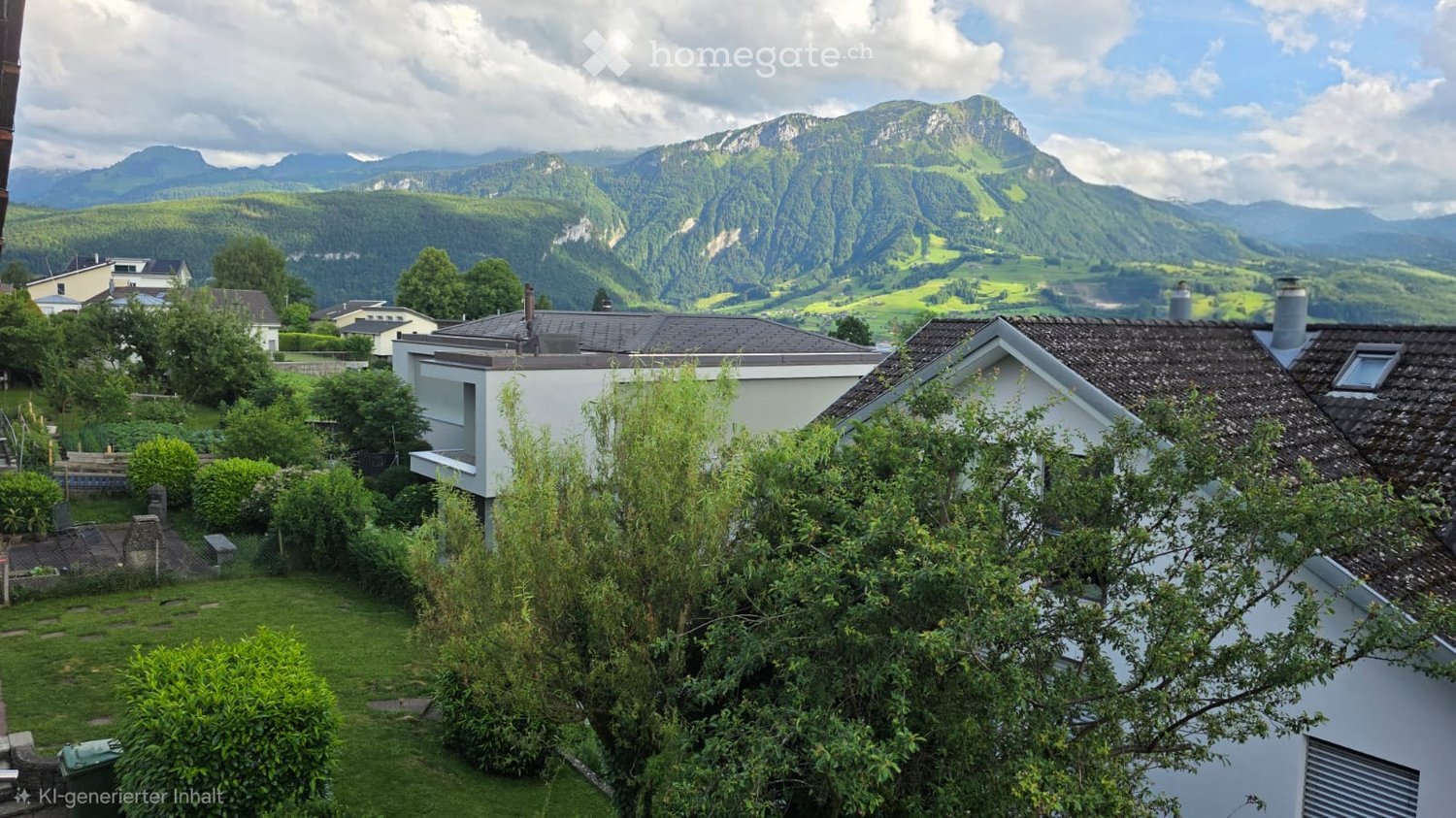 A serene garden view with a well-maintained lawn, various shrubs, and trees, overlooking a picturesque village with traditional houses and a stunning mountain backdrop.