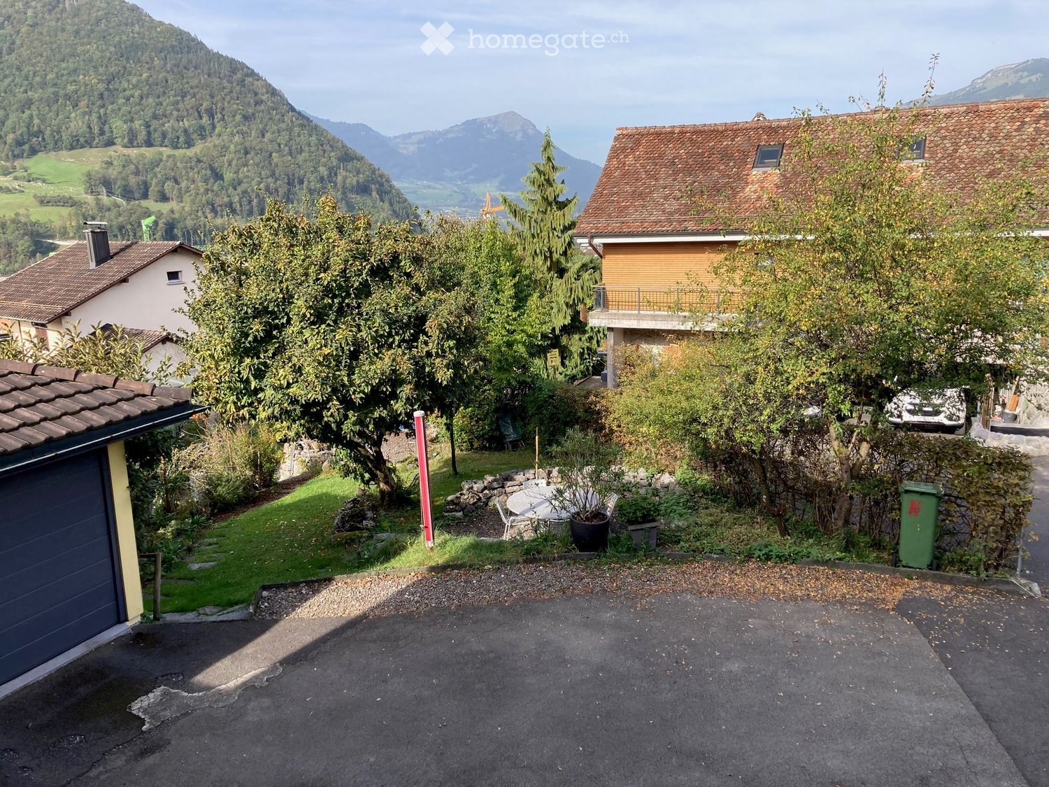 A serene garden area with a small patio, outdoor furniture, and a scenic mountain backdrop. The area includes a table, chairs, and a trash bin, surrounded by trees and greenery.
