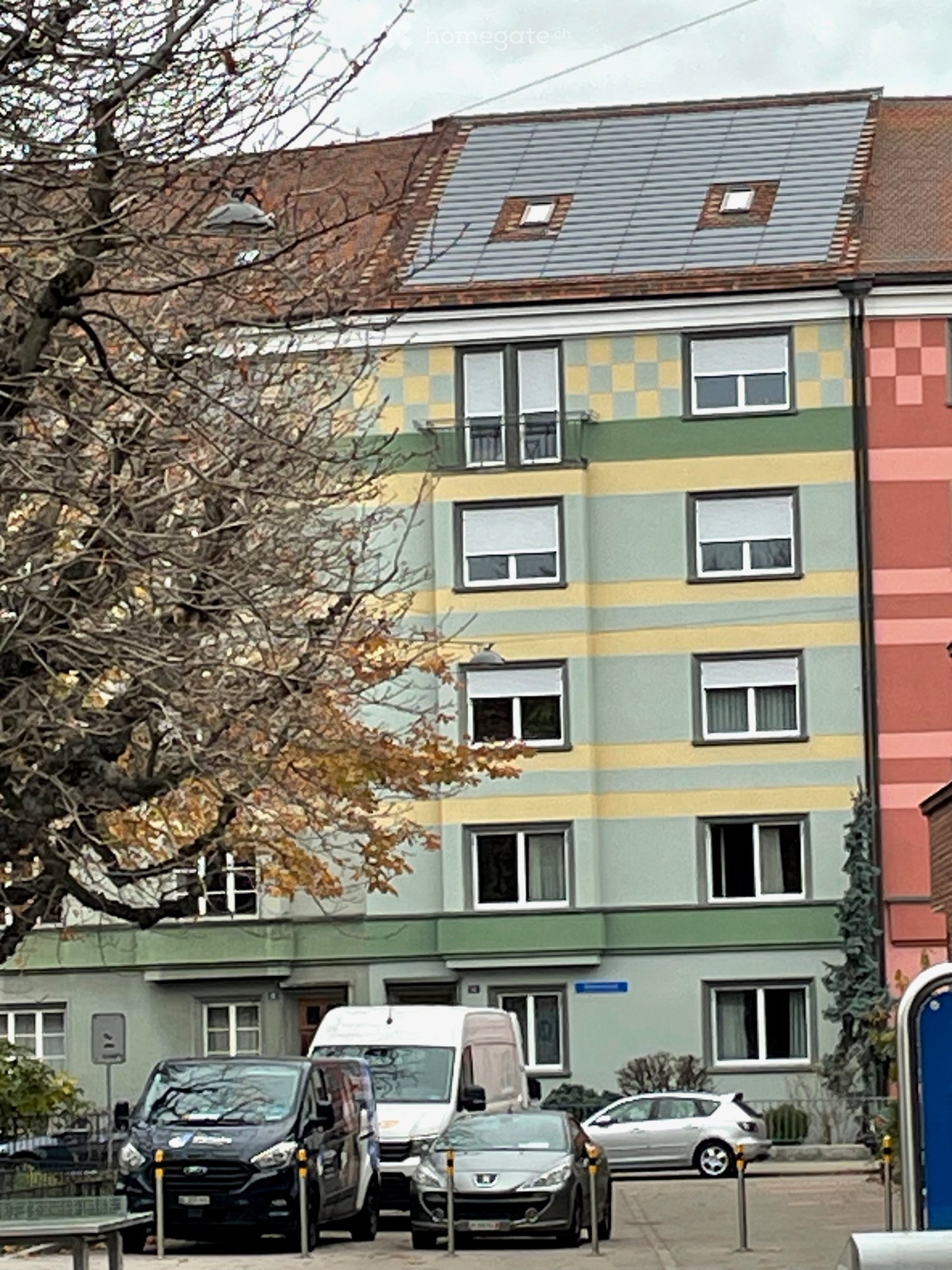 A two-story residential building with colorful striped facade, multiple windows, and a flat roof with skylights. Several cars are parked in front.