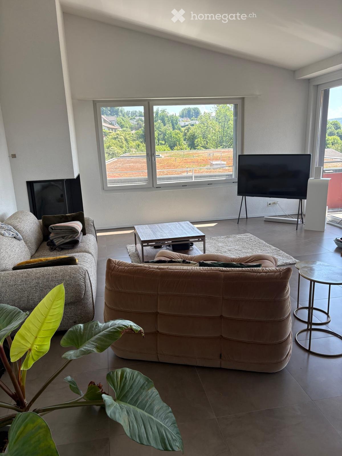 Modern living room with large windows offering a scenic view, featuring a grey sofa, a brown ottoman, a coffee table, a round side table, and a large screen.