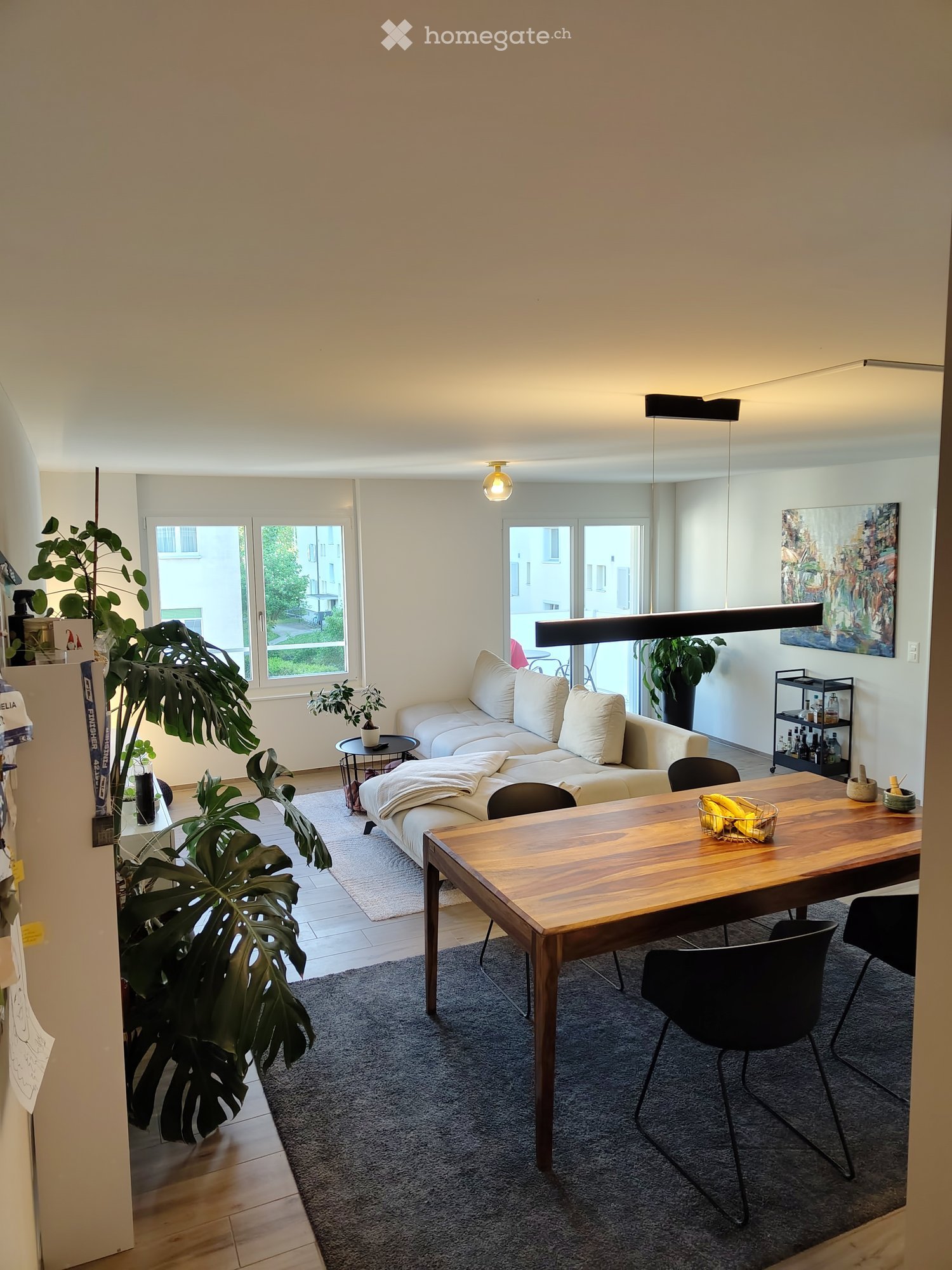 Furnished Room. Modern living room with a white sofa, wooden dining table, black chairs, potted plants, and a painting on the wall.