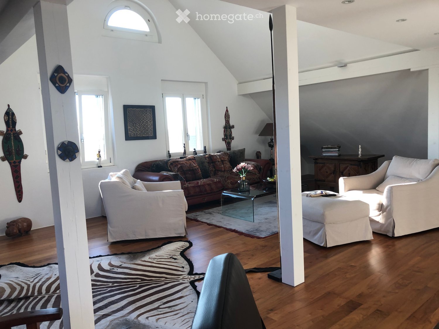 Bright living room with wooden flooring, white walls, large windows, and skylight. Features a cream-colored sofa, a brown leather sofa, a glass coffee table, a tall floor lamp, and decorative rugs.
