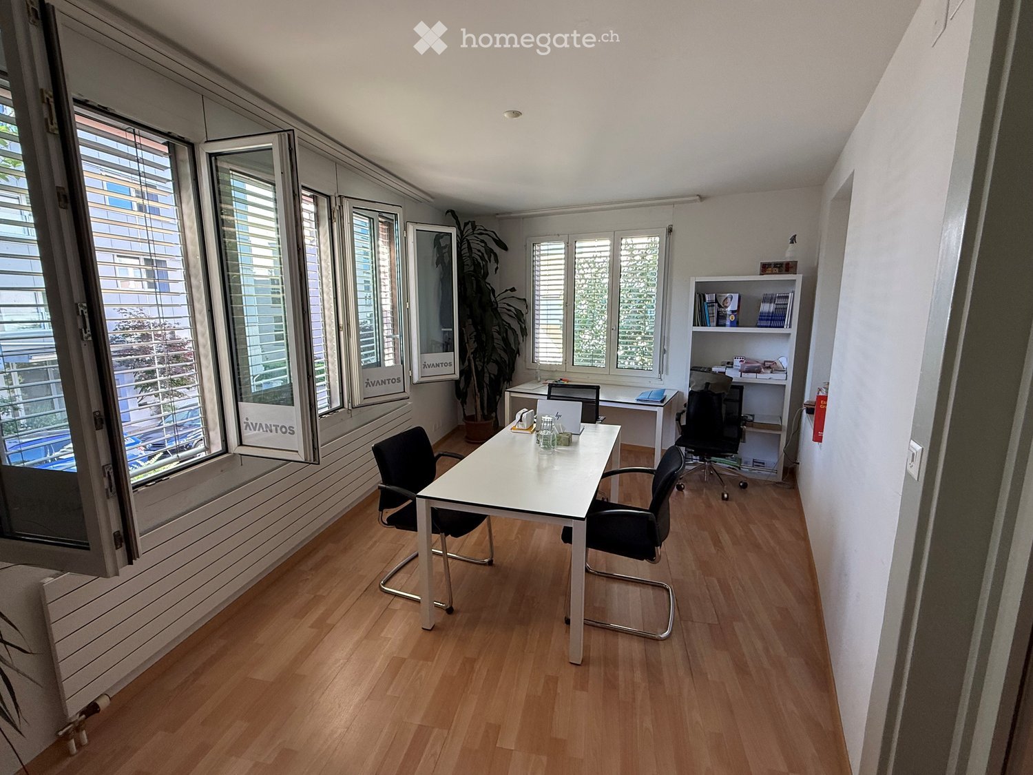 Modern office space with wooden flooring, large windows, a white desk, chairs, a laptop, and a bookshelf with books and decorative items.