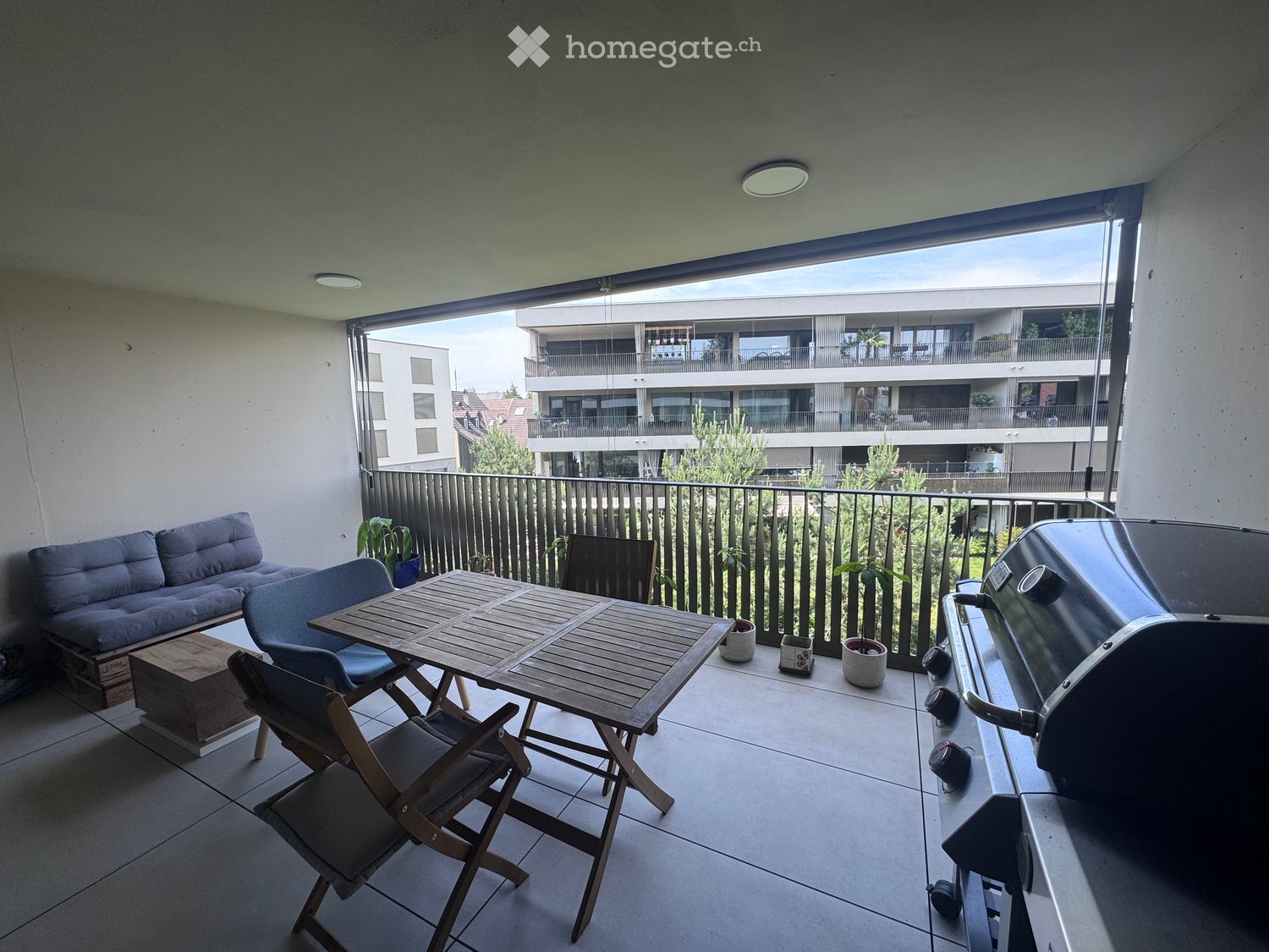 Balcony with a sofa, wooden table, chairs, grill, and potted plants, overlooking a residential area.