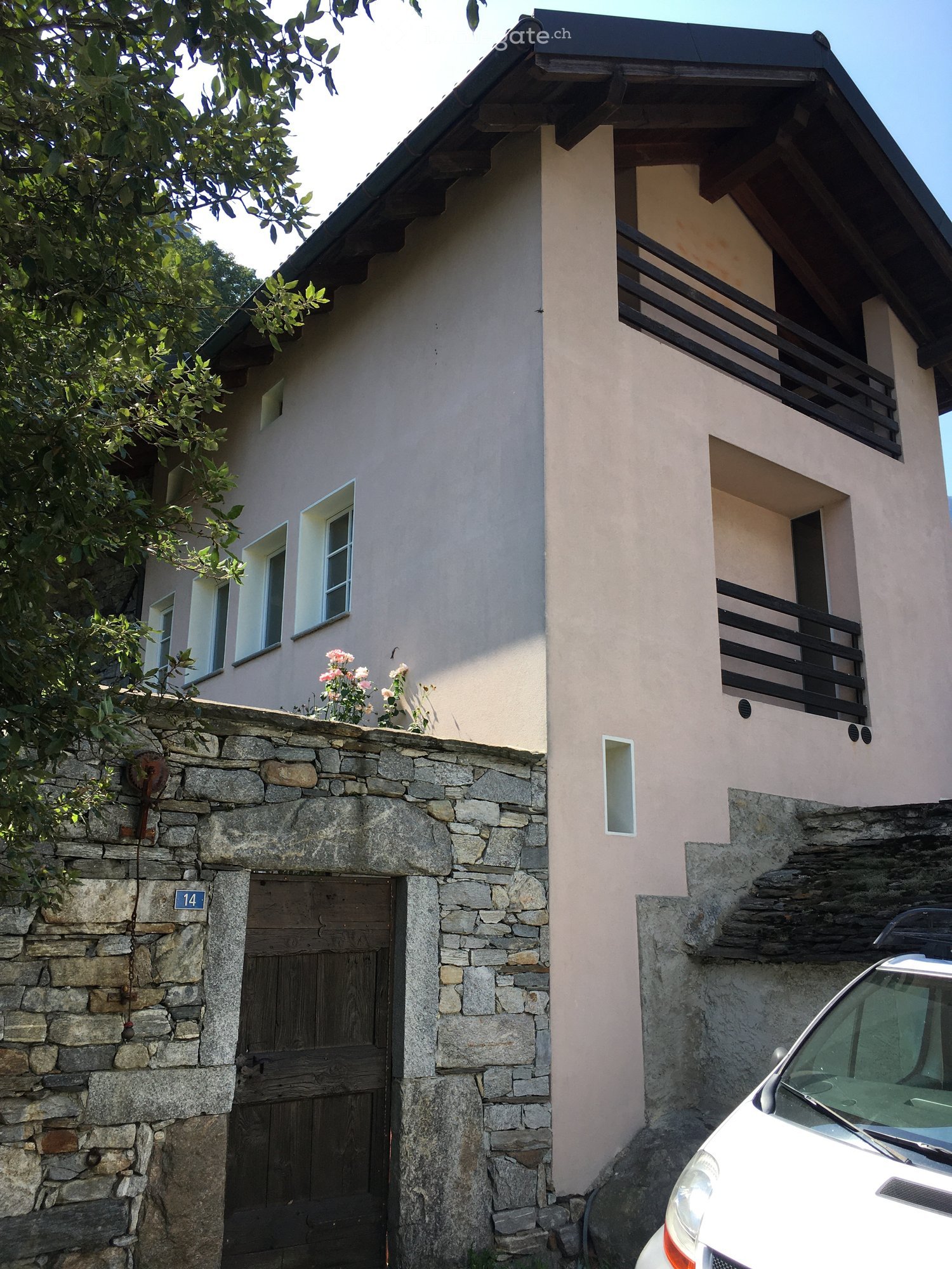 Two-story house with beige walls, black balcony railing, stone entrance, and a parked car in front.