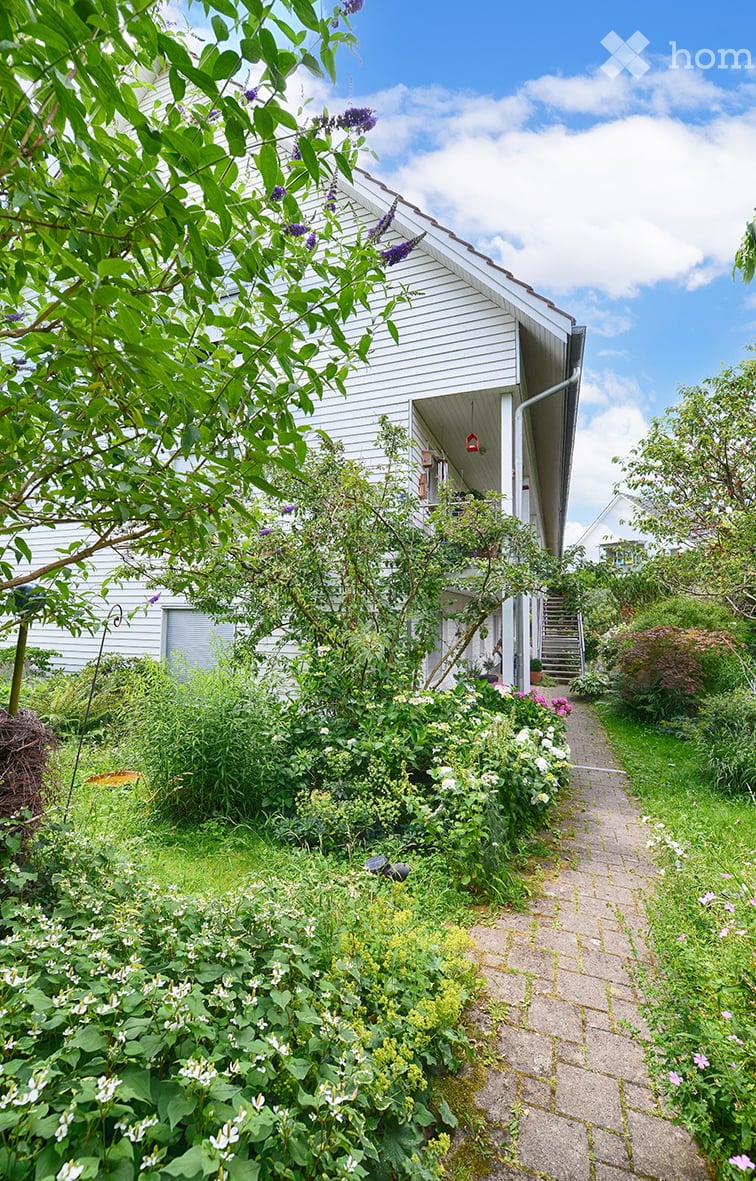 Two story house, white walls, wooden balcony, raised ground floor, brick pathway, plants, trees