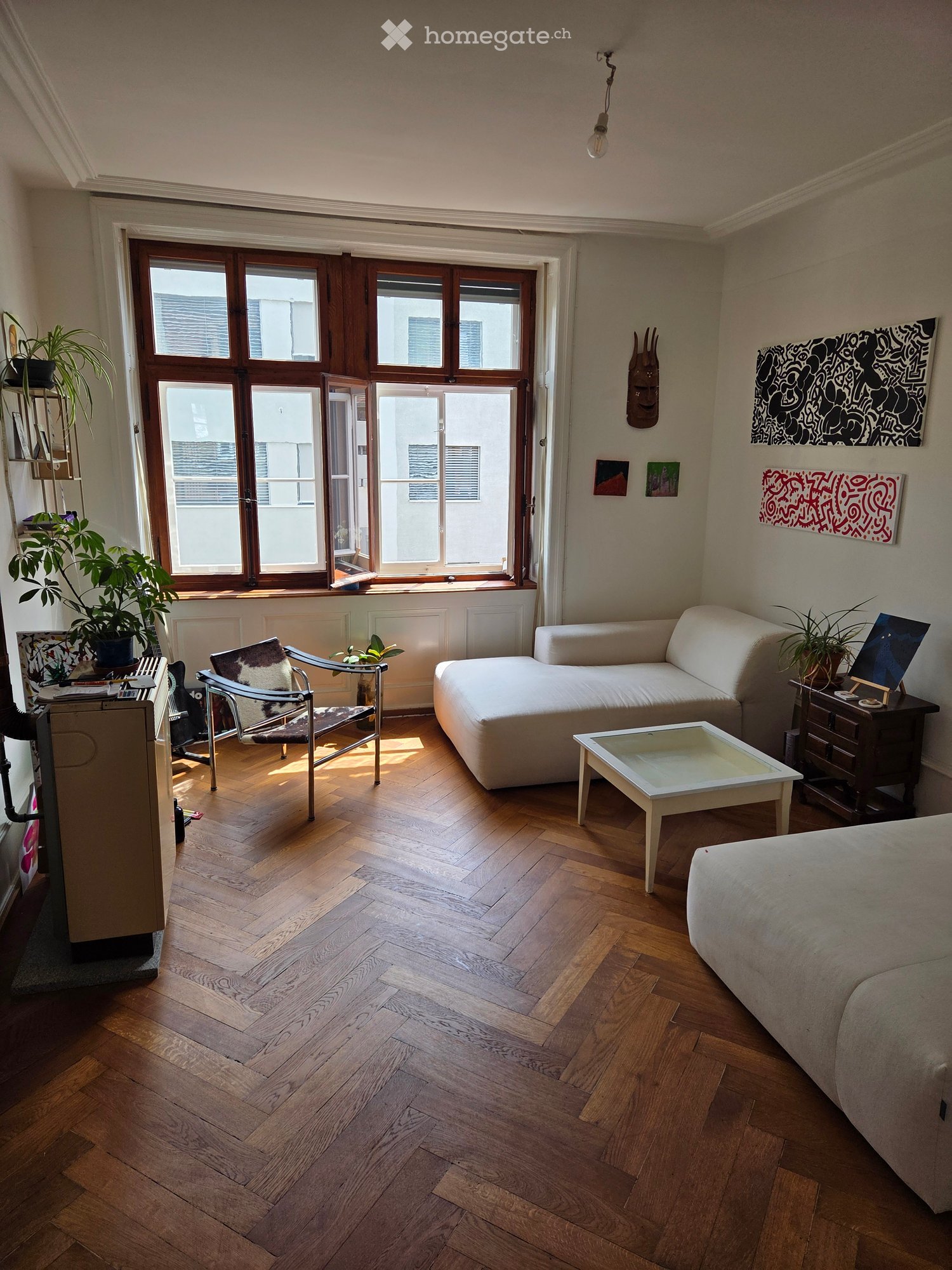 Modern living room with wooden flooring, large windows, white sofa, coffee table, and decorative art pieces.