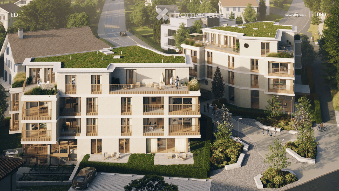 A modern multi-story residential building with balconies, green rooftop, and surrounding greenery. The building features large windows, wooden railings, and a paved parking area.