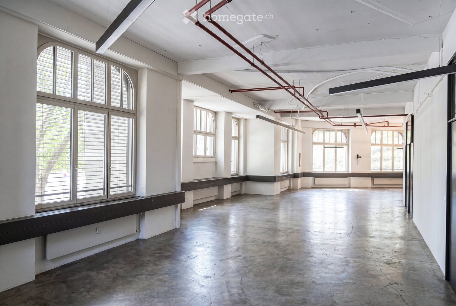Empty room with white walls, large windows, exposed pipes, and a concrete floor.