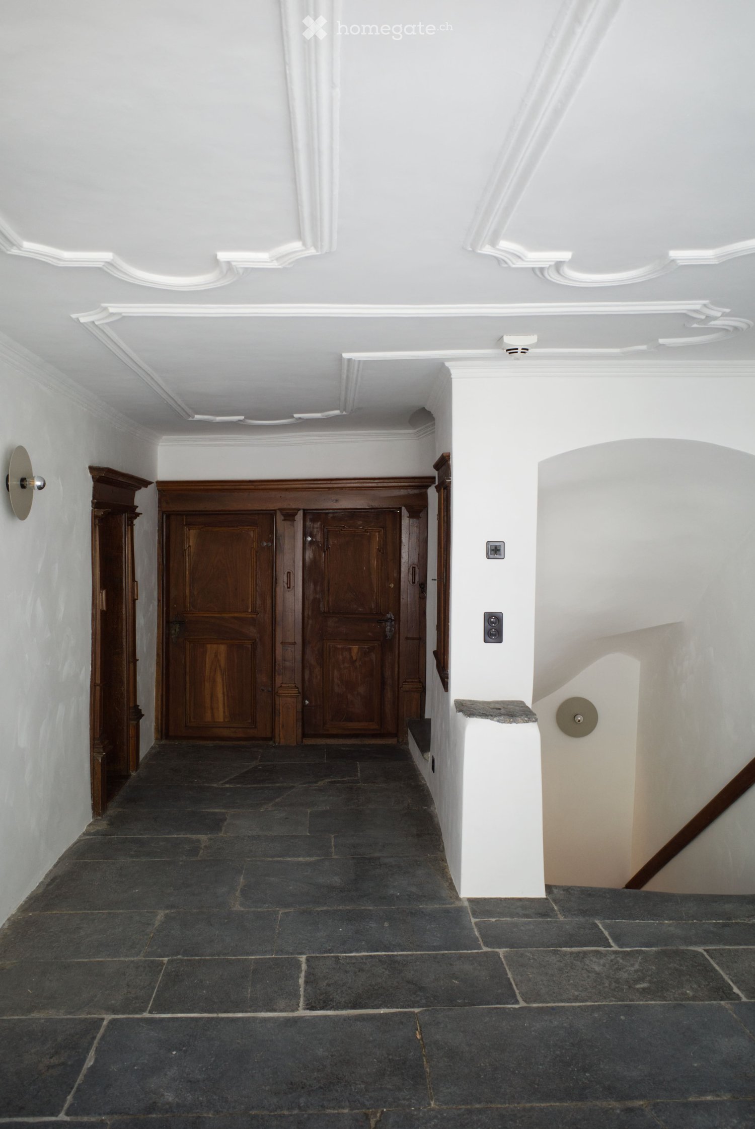 A long, narrow hallway with dark stone flooring, white walls, and ornate ceiling moldings. Features a wooden double door at the end and a small staircase on the right side.
