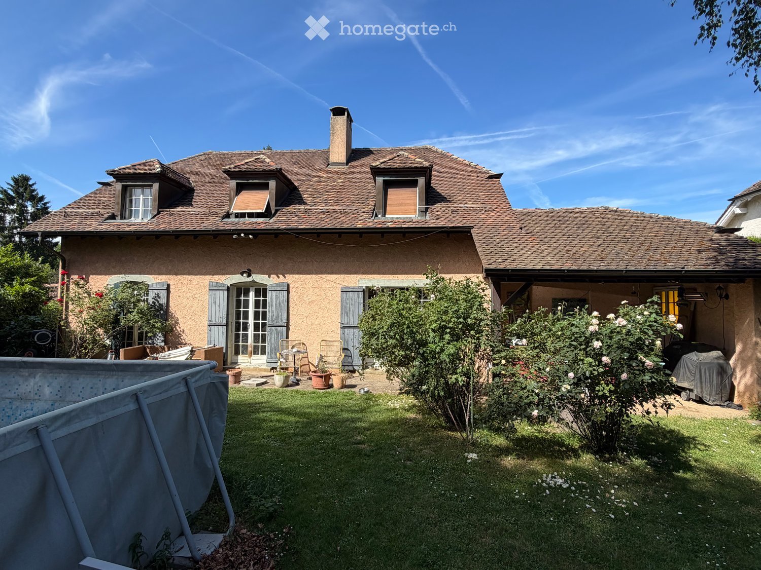 Two-story house with a tiled roof, multiple windows, and a chimney. The front yard features a lawn, a covered patio area, and several potted plants.