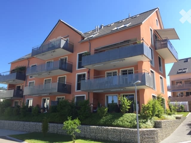 Modern multi-story apartment building with orange facade, multiple balconies, and greenery around the base.
