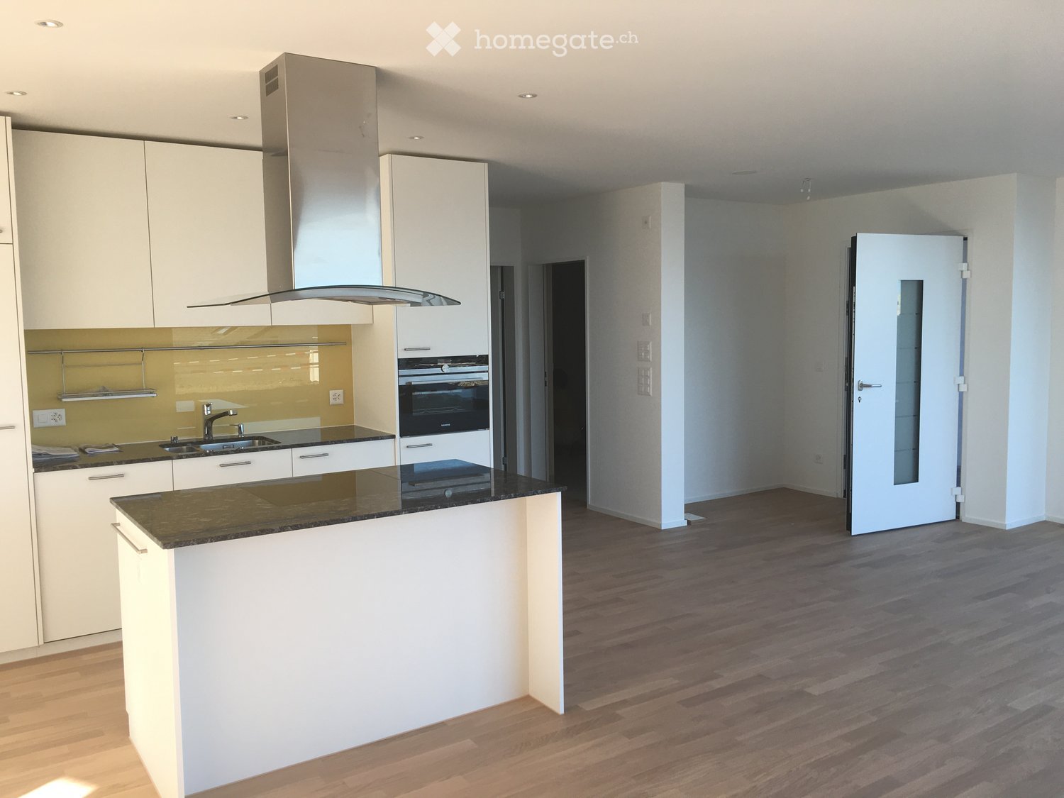 Modern kitchen with white cabinets, a central island, and a black countertop. Features include a sink, faucet, oven, and range hood. The floor is wooden, and the walls are light-colored.