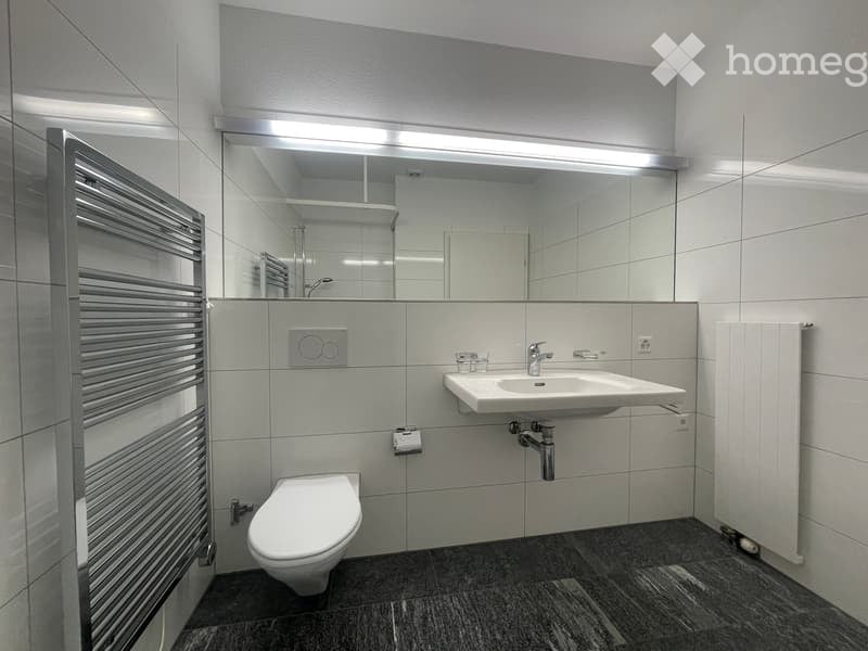 Modern bathroom with a large mirror, a white sink, a toilet, a radiator, and a sleek, dark tiled floor.