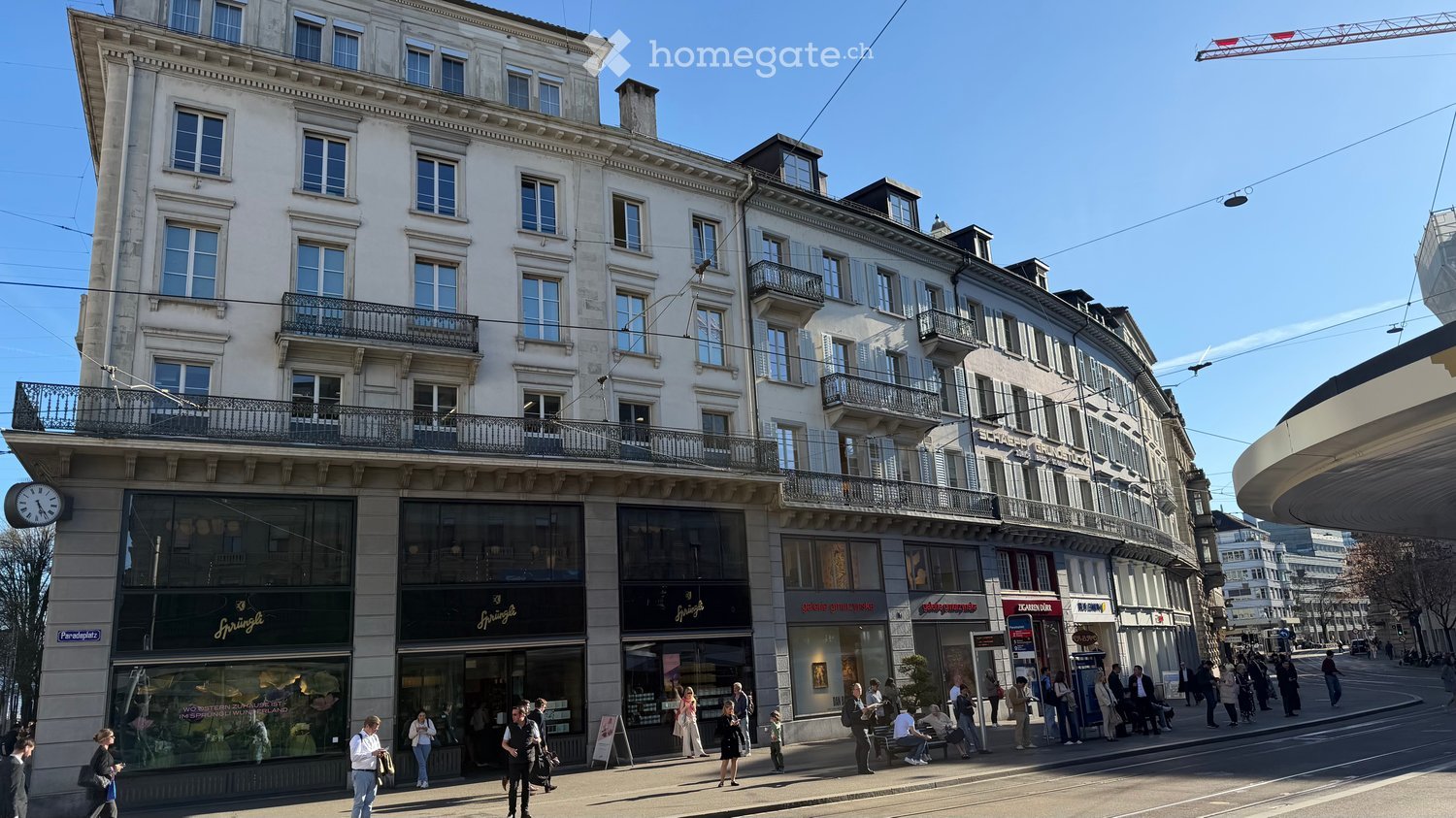Multi-story building with large windows, balconies, and storefronts. The ground floor features a shop with glass windows and signage, while pedestrians walk along the sidewalk.