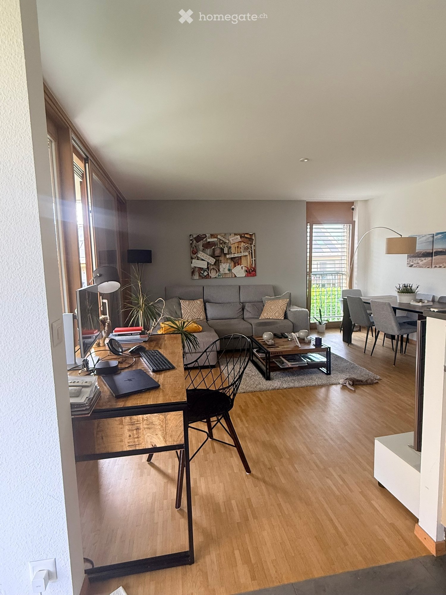 Modern living room with wooden flooring, grey sofa, coffee table, laptop, and dining area with chairs and table.