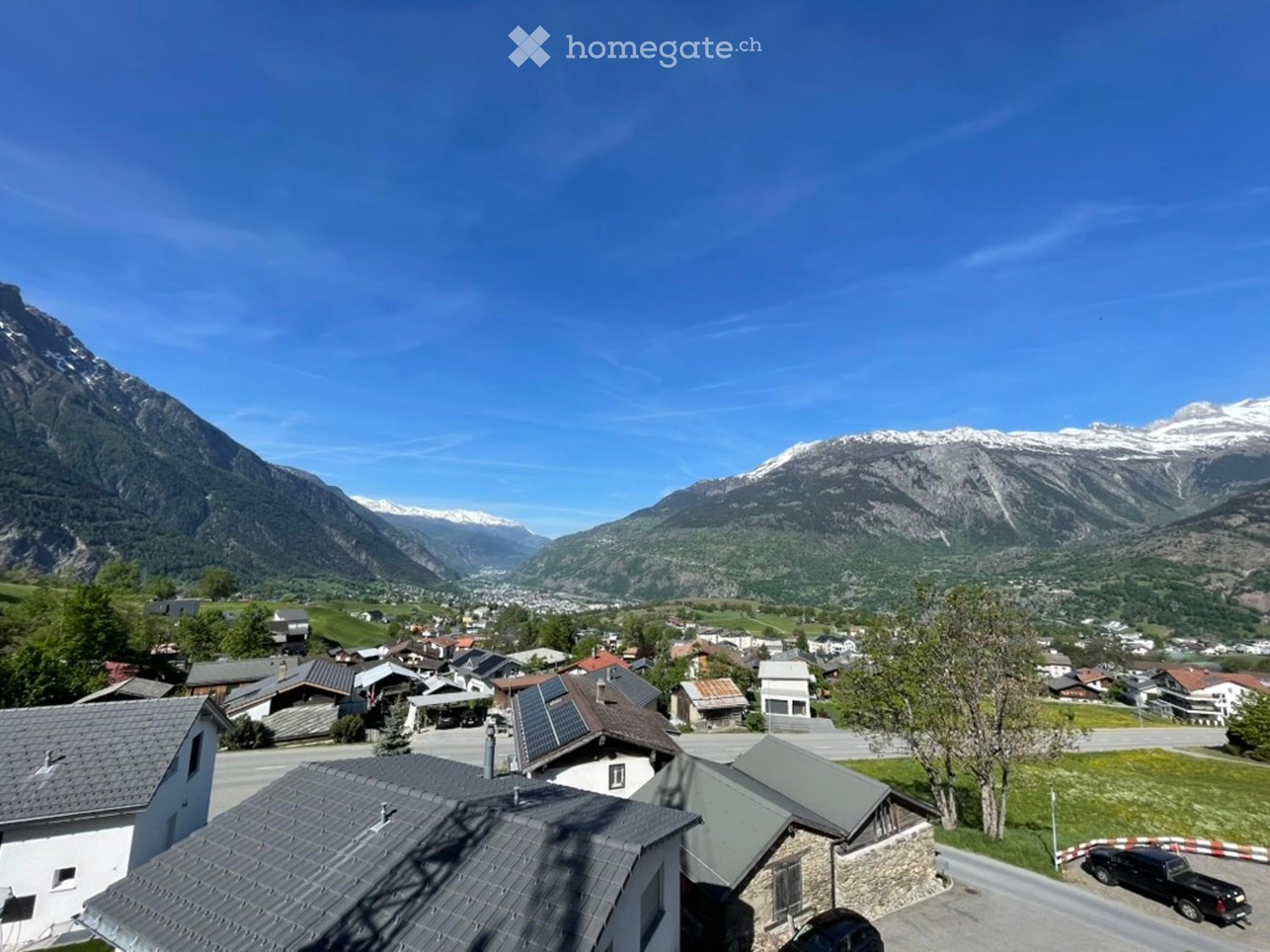 A panoramic view of a village nestled in a valley surrounded by mountains, with houses, roads, and a clear blue sky.