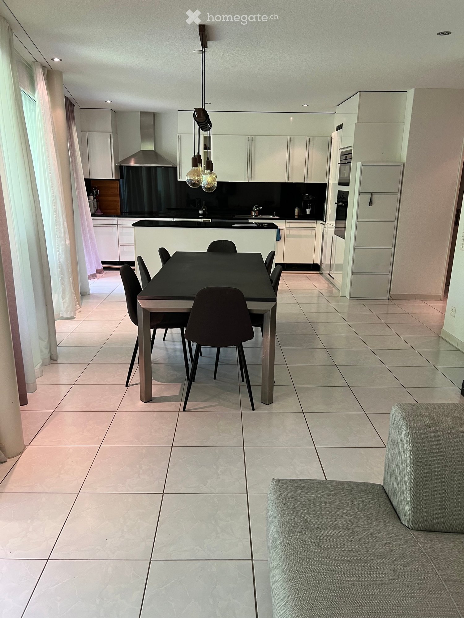 Modern kitchen and living area with white cabinets, a black dining table with six chairs, a grey sofa, and built-in appliances including a refrigerator and oven.