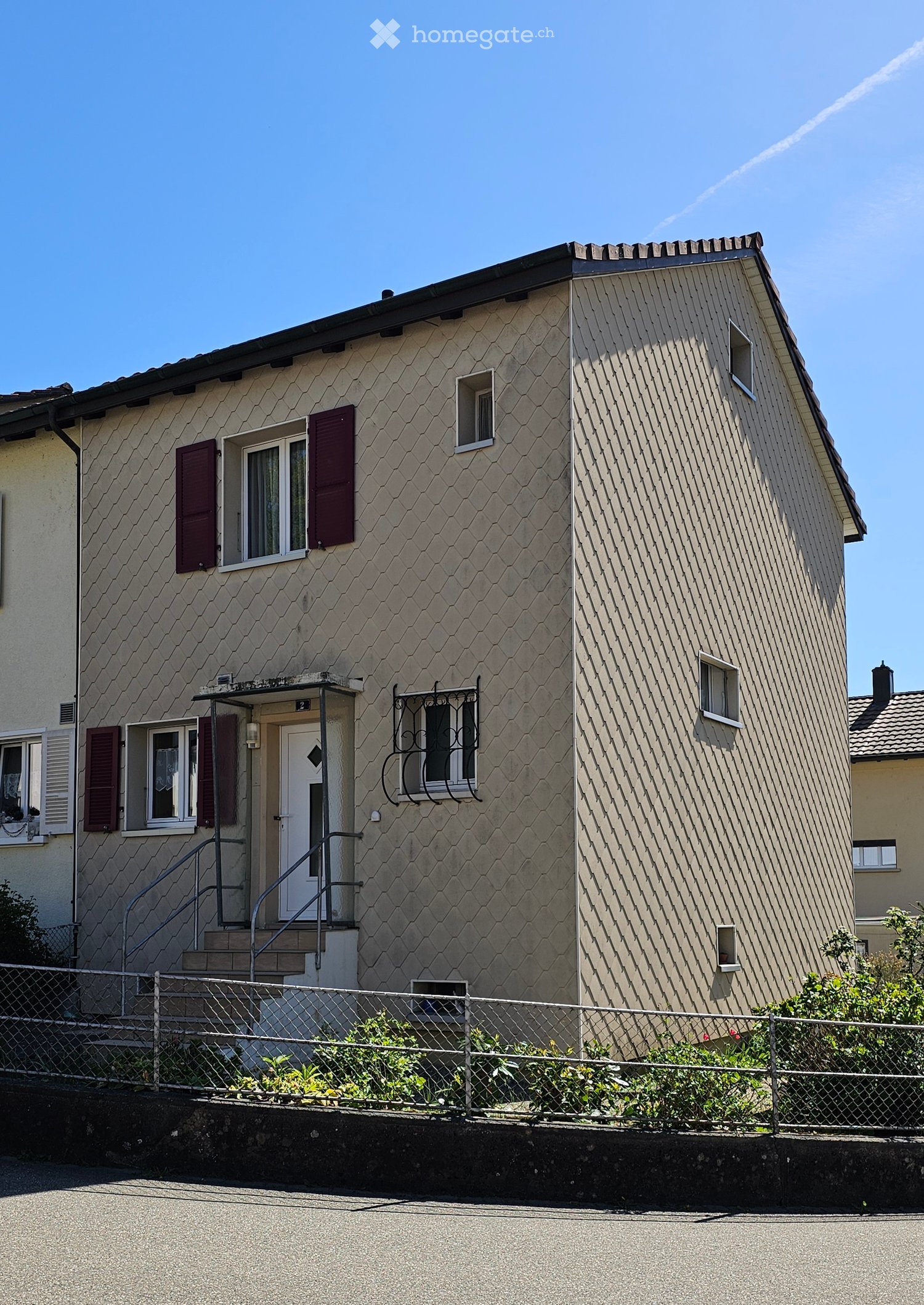 Two-story house with beige textured exterior, multiple windows with shutters, and a small staircase leading to the entrance.