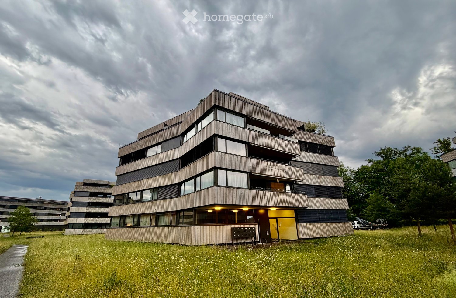 Modern multi-story building with wooden facade, large windows, and balconies, surrounded by green grass and trees.