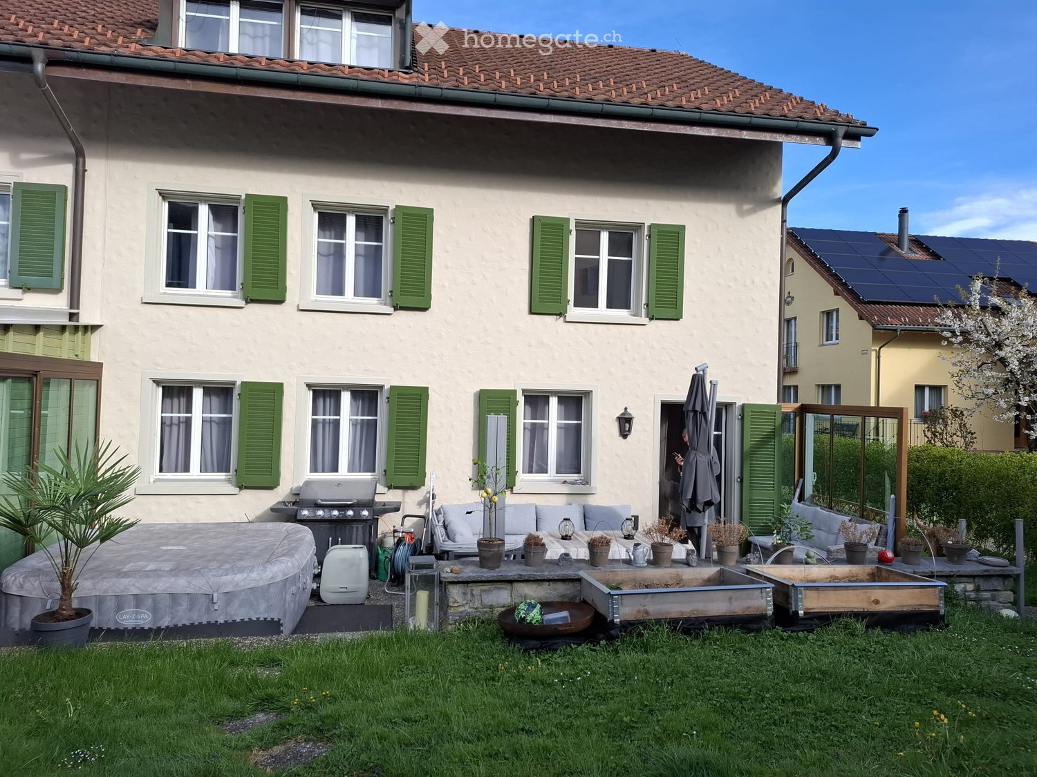 Garden / Outdoor Area. A well-maintained garden area with a hot tub, sofa, table, grill, potted plants, and an umbrella. The house in the background has green shutters and multiple windows.