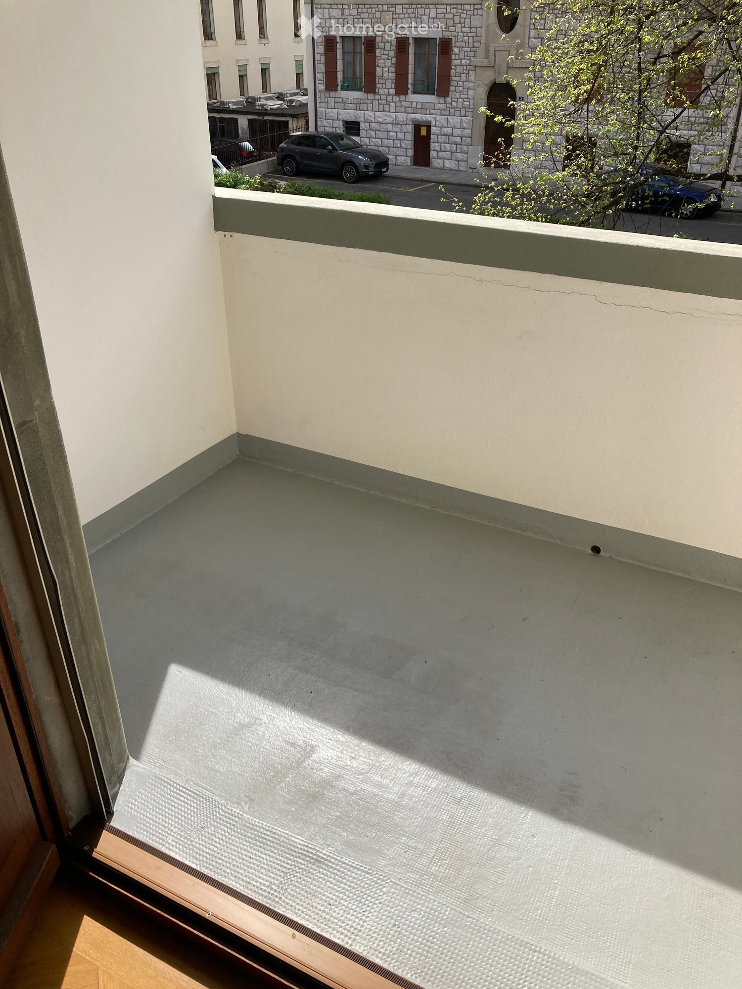 Balcony with concrete floor, white walls, and a view of a street with parked cars and a building with stone facade and shutters.