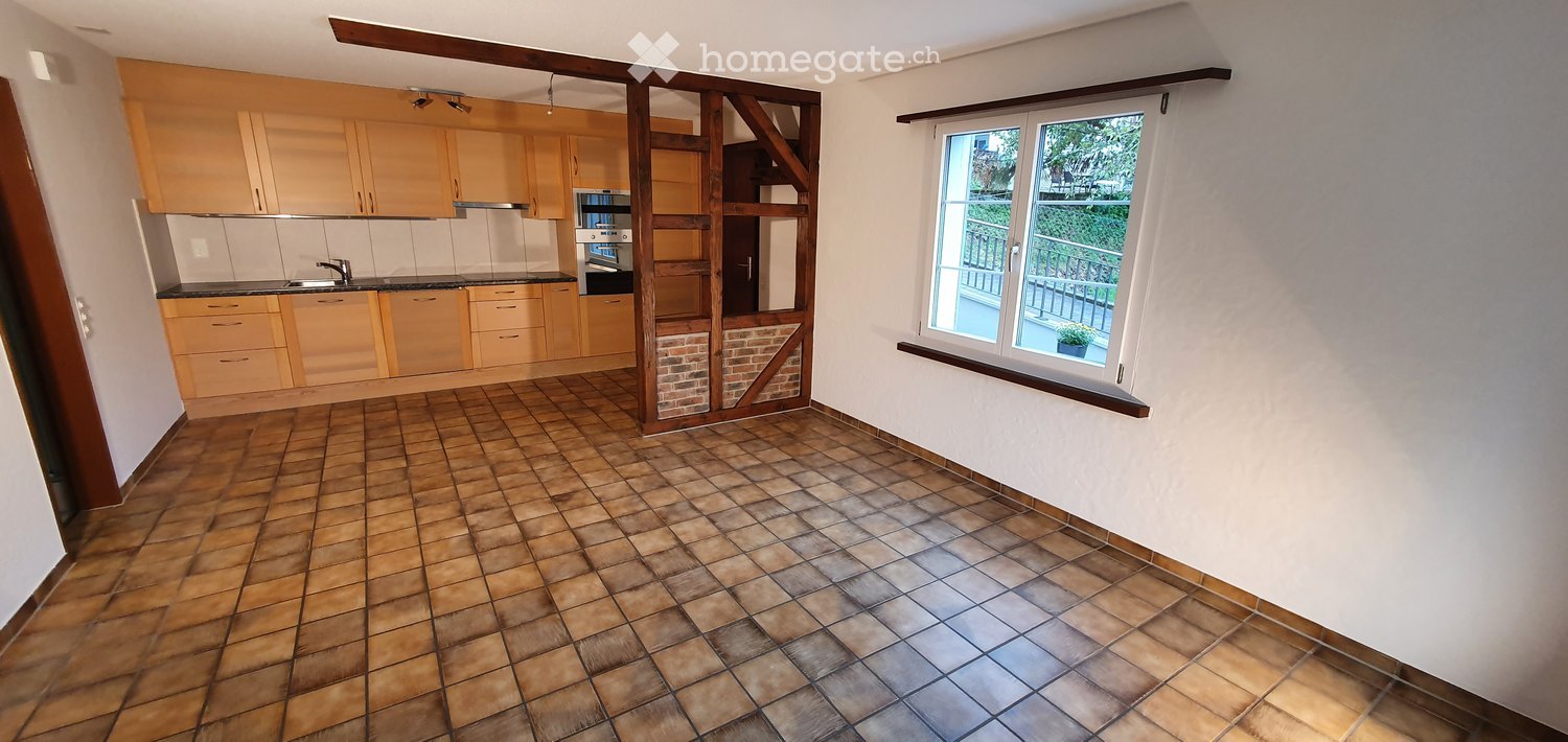 The image shows a modern kitchen with light wooden cabinets, a black countertop, and a sink with a faucet. There is also an oven and a microwave visible. The floor is tiled in a warm, earthy tone, and the room has large windows with a view of a balcony.