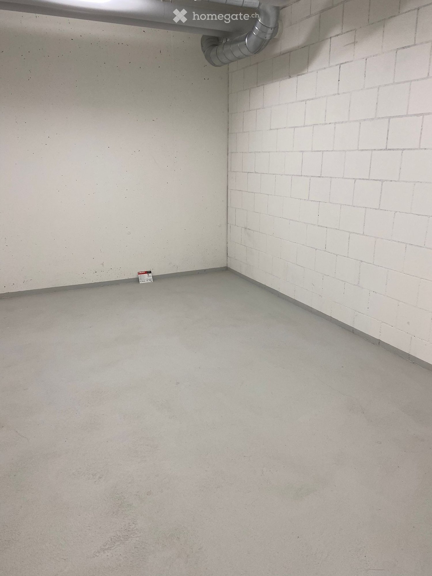 Empty room with concrete floor and white tiled wall, featuring a metal pipe and a small sign on the floor.